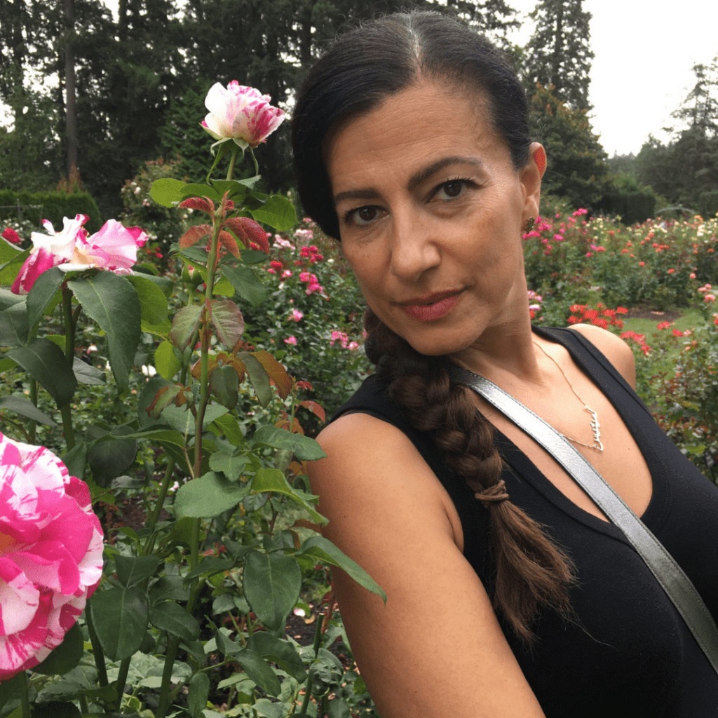 A middle aged olive skinned woman with dark hair in a braid. She is standing next to a rose bush of striped pink and white roses.